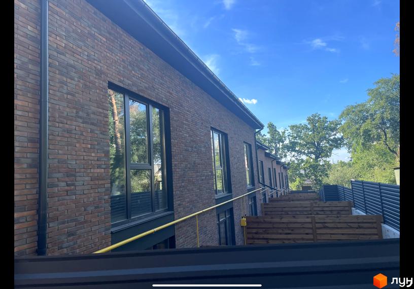 Таунхауси „Сингаївського“: цегляні фасади будинків з великими вікнами та дерев'яні тераси на тлі зелених дерев та блакитного неба.