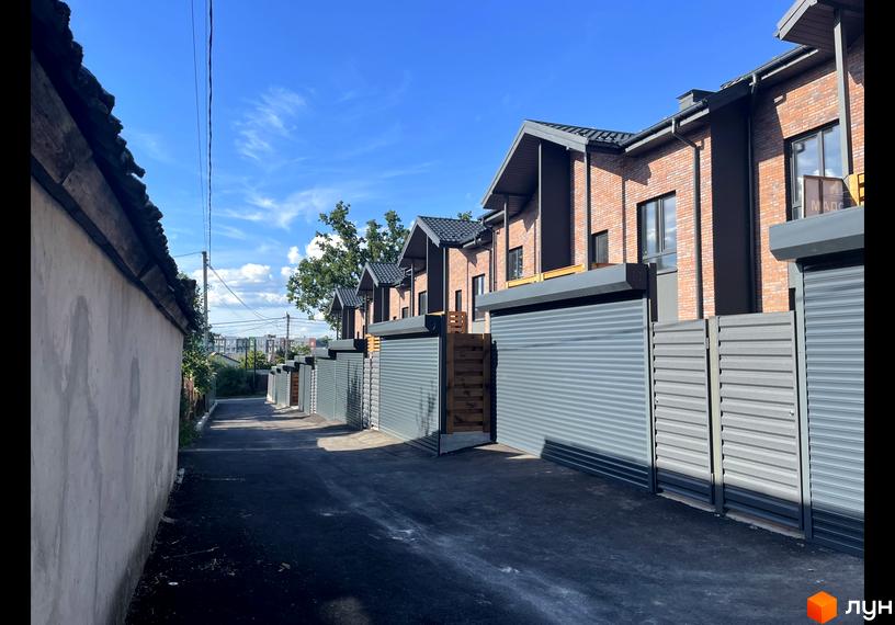 Таунхауси Сингаївського: цегляні будинки з сірими ролетами та парканами, асфальтована дорога та зелені насадження під блакитним небом.