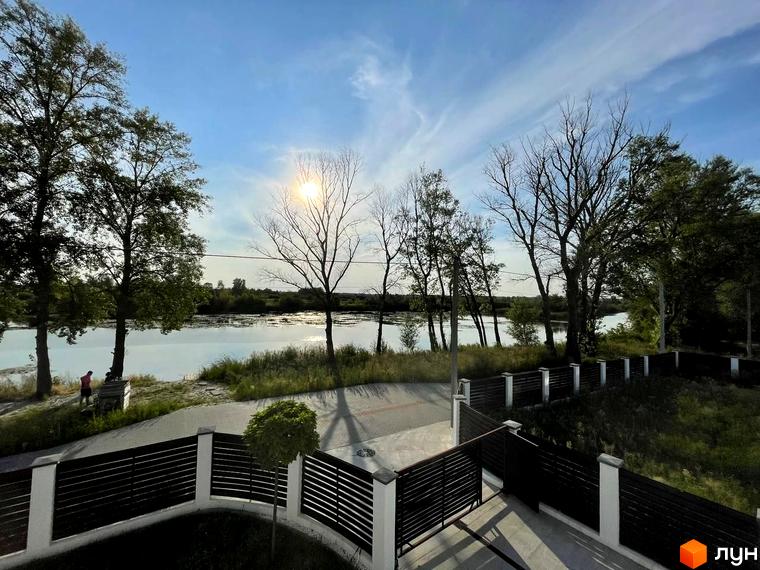 Котеджне містечко Sunrise Residence з видом на річку та зелені насадження. Територія огороджена парканом з білими стовпами та темними ламелями.