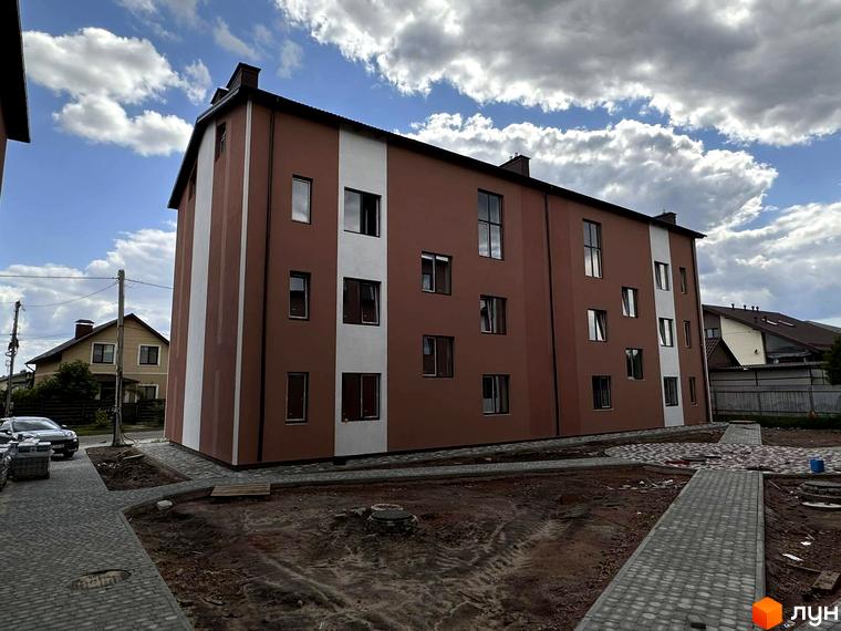 Зовнішній вигляд будинку житлового комплексу Riviera з коричневим фасадом та білою смугою, прибудинкова територія з бруківкою та зеленими насадженнями.