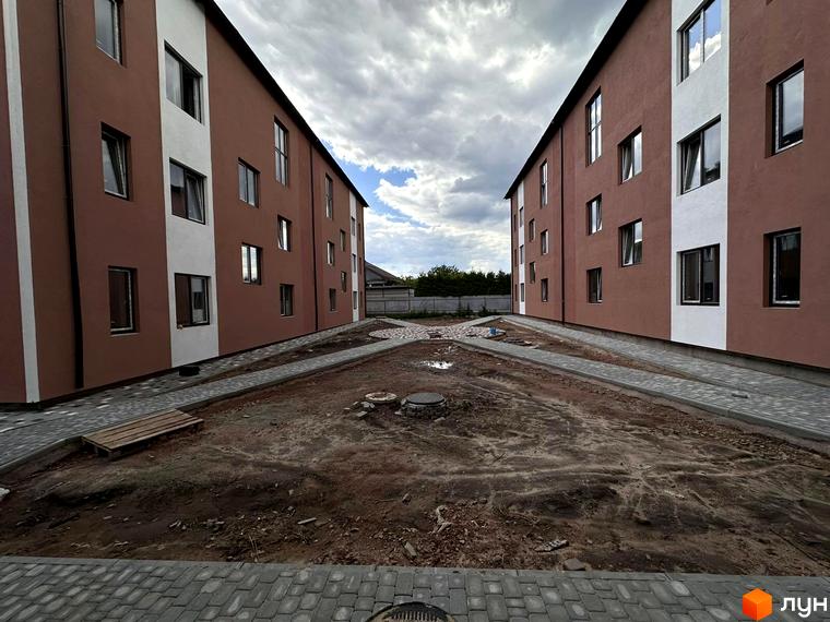 Зовнішній вигляд будинків житлового комплексу Riviera з коричневим та білим фасадом. На прибудинковій території видно бруківку та ділянку з землею.