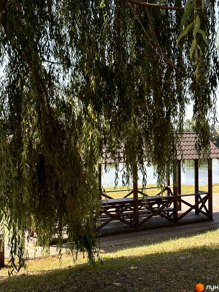 Альтанка з дерев'яними столами та лавками біля річки, оточена плакучими вербами. Територія біля новобудови ЖК Riviera з зеленою травою та сонячними променями.
