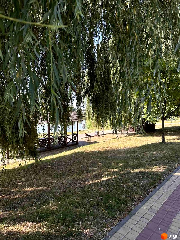 Житловий комплекс Riviera біля озера з альтанкою та місцями для відпочинку. Зелена зона з деревами та доріжкою для прогулянок.