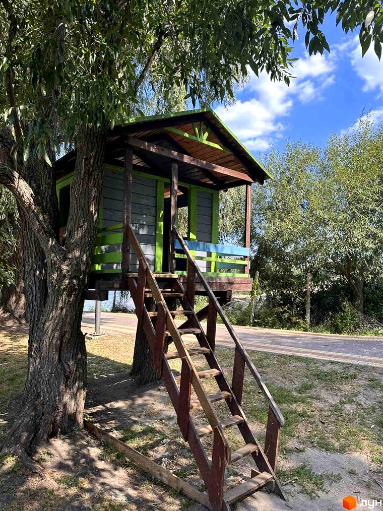 Дитячий будиночок на дереві в ЖК Riviera з дерев'яними сходами та зеленою огорожею. Навколо зелені дерева та блакитне небо з хмарами.