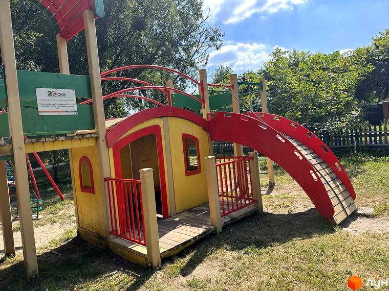 Дитячий майданчик у житловому комплексі Riviera з яскравою жовтою гіркою та червоними перилами. Навколо майданчика зелена трава та дерева.