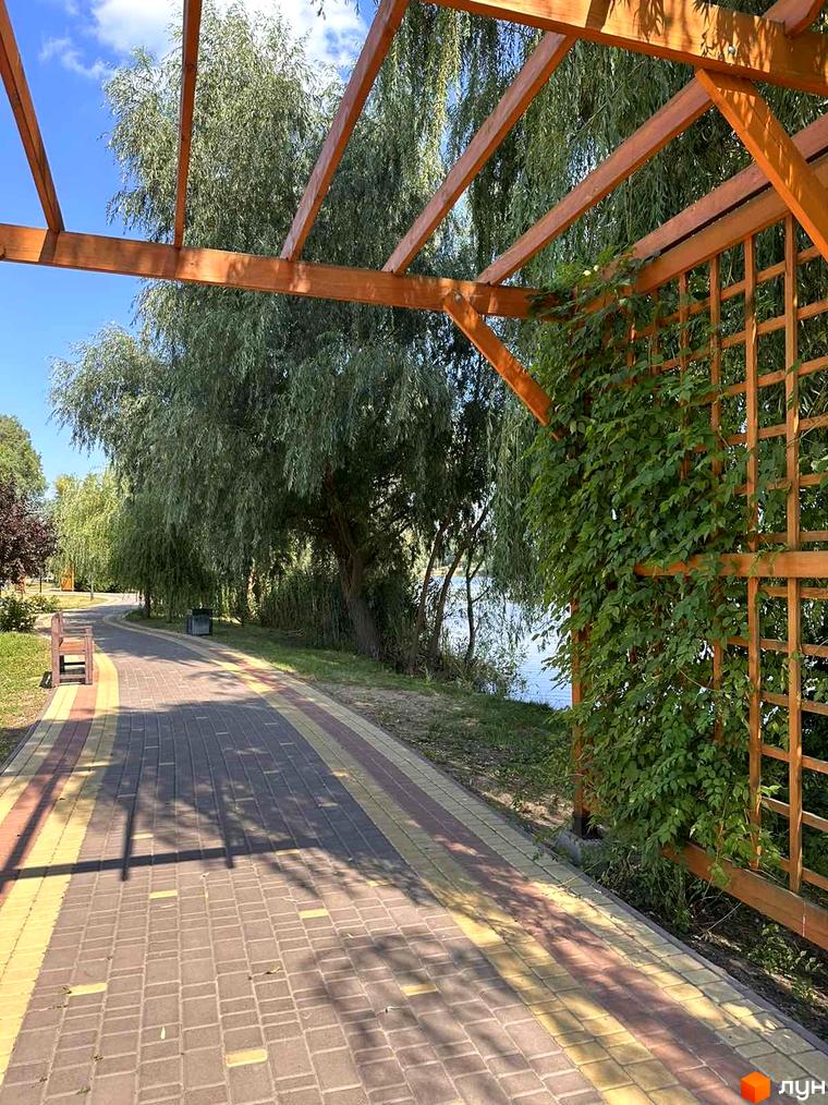 Житловий комплекс Riviera: дерев'яна альтанка з ліанами та доріжка з бруківки біля озера. Навколо зелені дерева та кущі.