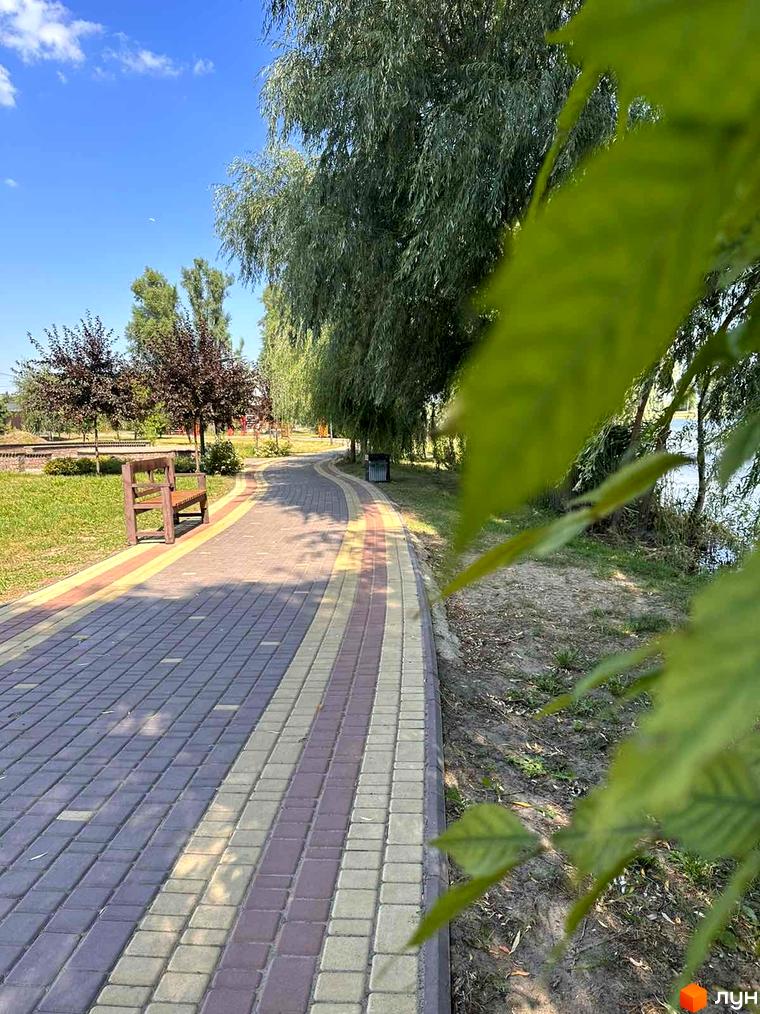 Житловий комплекс Riviera: мальовнича набережна з пішохідною доріжкою та лавкою для відпочинку. Зелені дерева та вода створюють затишну атмосферу.