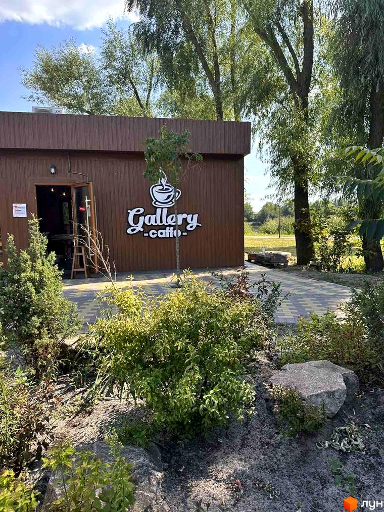 Зовнішній вигляд кафе Gallery Caffe з коричневими стінами та вхідними дверима. Навколо кафе розташована зелена зона з деревами та кущами.