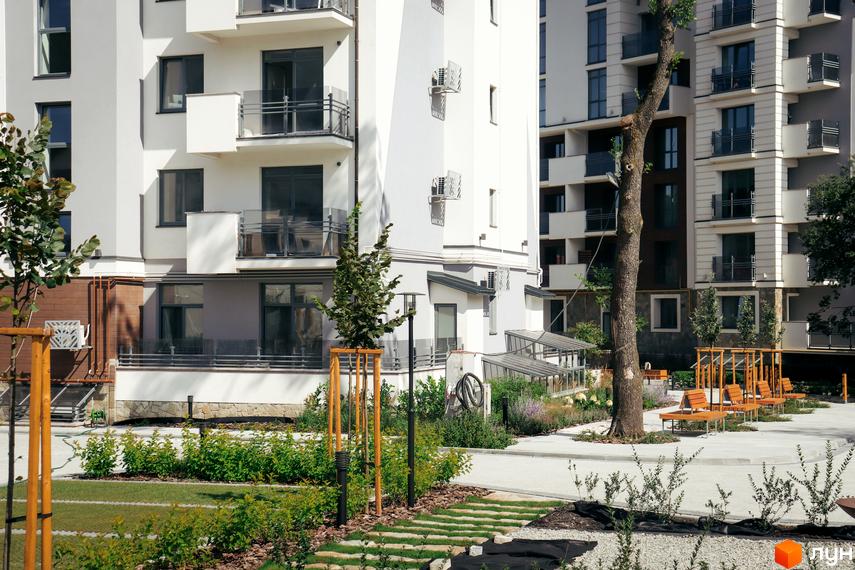 Новобудова Крайобраз з білими фасадами, скляними балконами та доглянутою прибудинковою територією з деревами та зонами відпочинку.