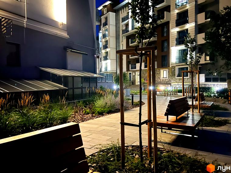 Житловий комплекс Крайобраз: фасад будинку та освітлена прибудинкова територія з лавками та деревами ввечері.