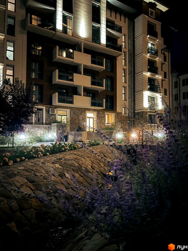 Житловий комплекс Крайобраз вночі. Зовнішній вигляд будівлі з підсвіченими вікнами та балконами, кам'яною облицовкою та ландшафтним дизайном прибудинкової території з квітами та освітленням.