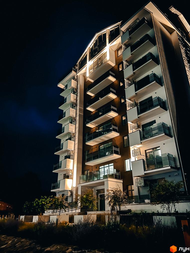 Житловий комплекс Крайобраз вночі. Багатоквартирний будинок з балконами та підсвіткою. Навколо будинку зелені насадження та освітлення.