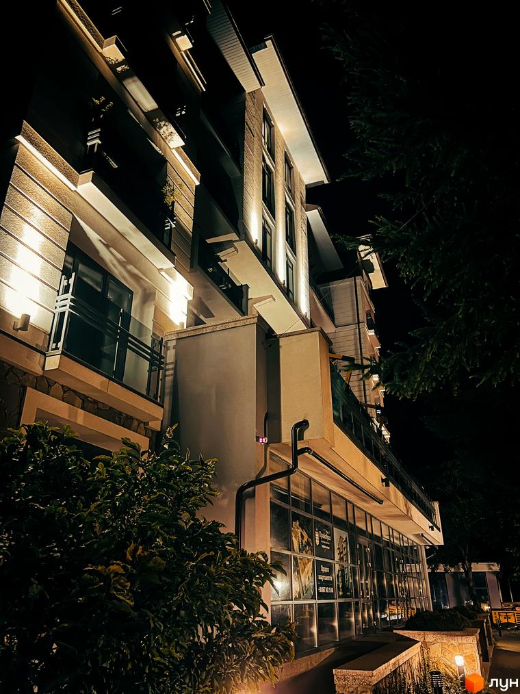 Житловий комплекс Крайобраз вночі. Зовнішній вигляд будівлі з підсвіченими фасадами та вікнами. Прибудинкова територія з деревами та освітленням.