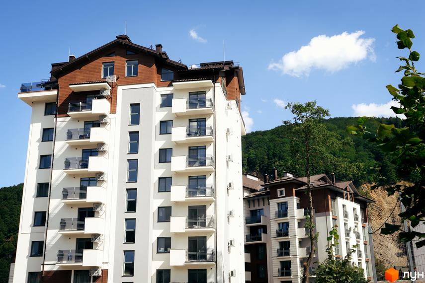 Зовнішній вигляд новобудови Крайобраз з білим фасадом та коричневими елементами, балконами та вікнами на тлі зеленого гірського схилу. Фотографія житлового комплексу в сонячний день.