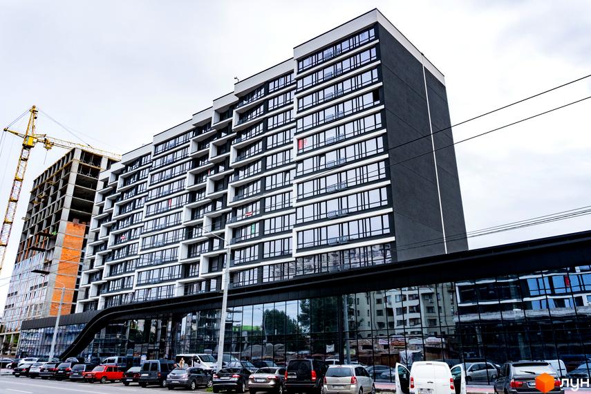 Зовнішній вигляд новобудови ЖК А5 з великою кількістю вікон та балконів. Перед будинком розташована парковка з автомобілями.