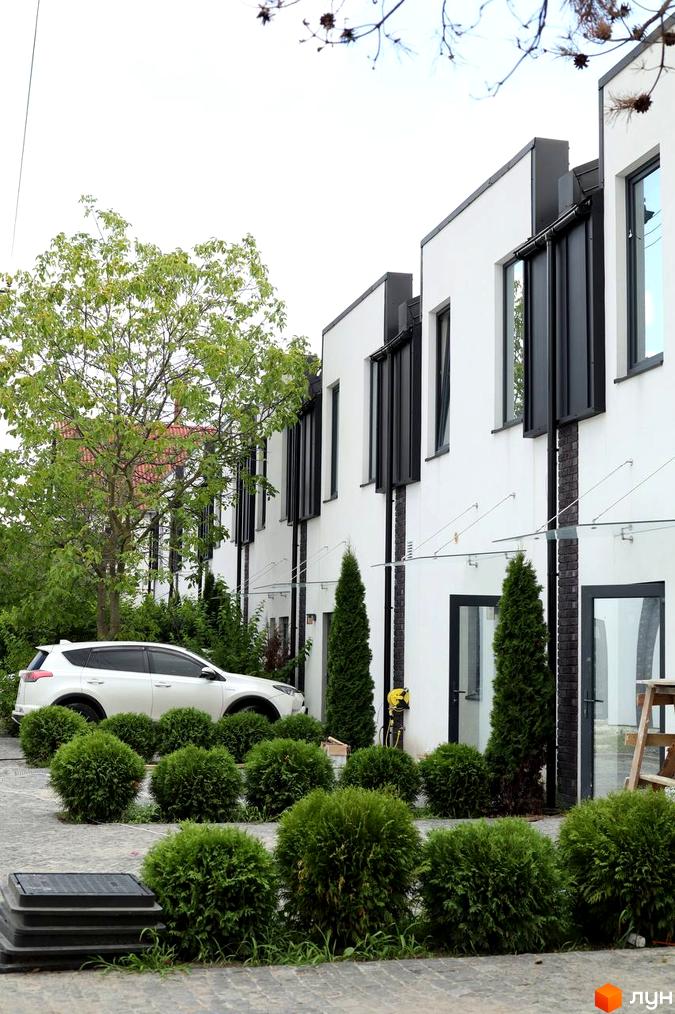 Зовнішній вигляд таунхаусів Lisostepoville з білими фасадами та чорними вікнами, прибудинкова територія з зеленими кущами та автомобілем.