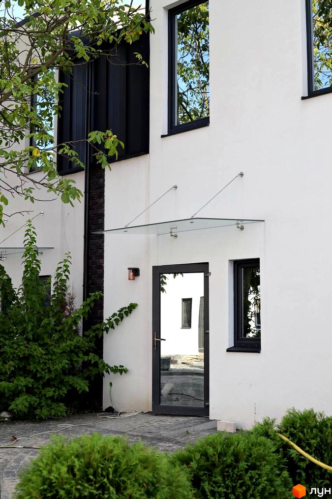 Зовнішній вигляд таунхаусів Lisostepoville з білою стіною, скляним навісом над дверима та зеленими насадженнями навколо. Сучасна архітектура новобудови з великими вікнами.