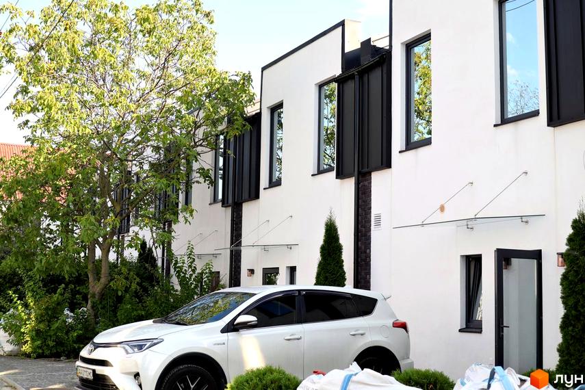 Зовнішній вигляд таунхаусів Lisostepoville з білими стінами та чорними вікнами. На передньому плані автомобіль та зелені насадження.