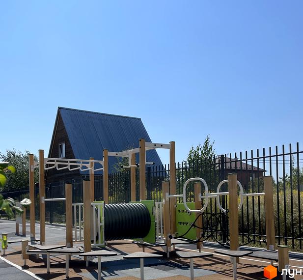 Житловий комплекс Art Misto: дитячий майданчик з елементами для спорту на тлі дерев'яного будинку під синім небом.