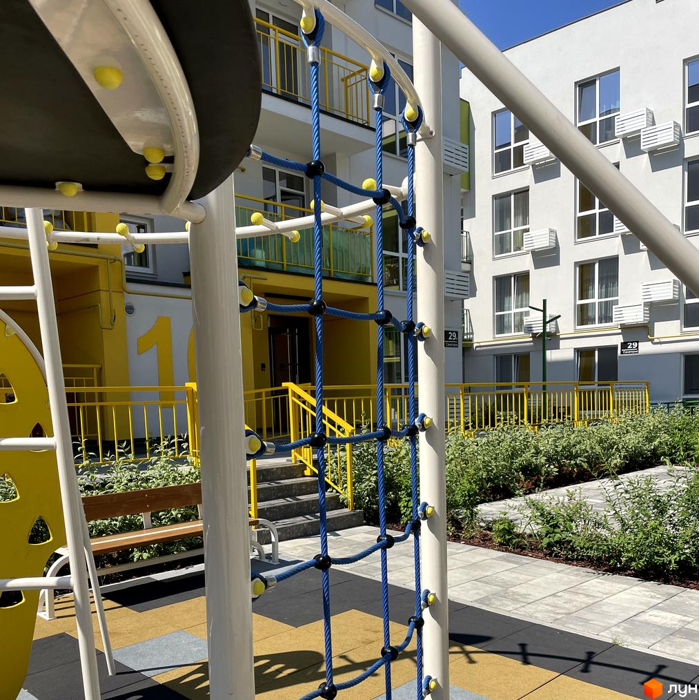 Житловий комплекс Art Misto: білі фасади будинків з кондиціонерами та жовті огорожі. Поруч дитячий майданчик з синіми канатами та зелені насадження.