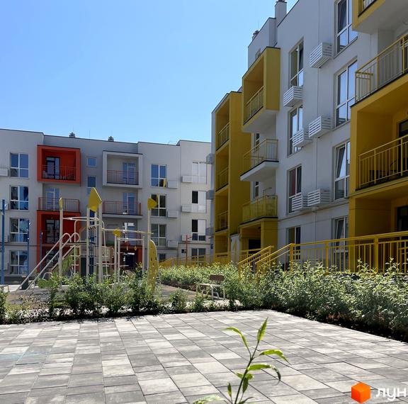Житловий комплекс Art Misto з яскравими жовтими та помаранчевими елементами фасадів та дитячим майданчиком на тлі блакитного неба. Територія комплексу упорядкована, викладена плиткою та озеленена.