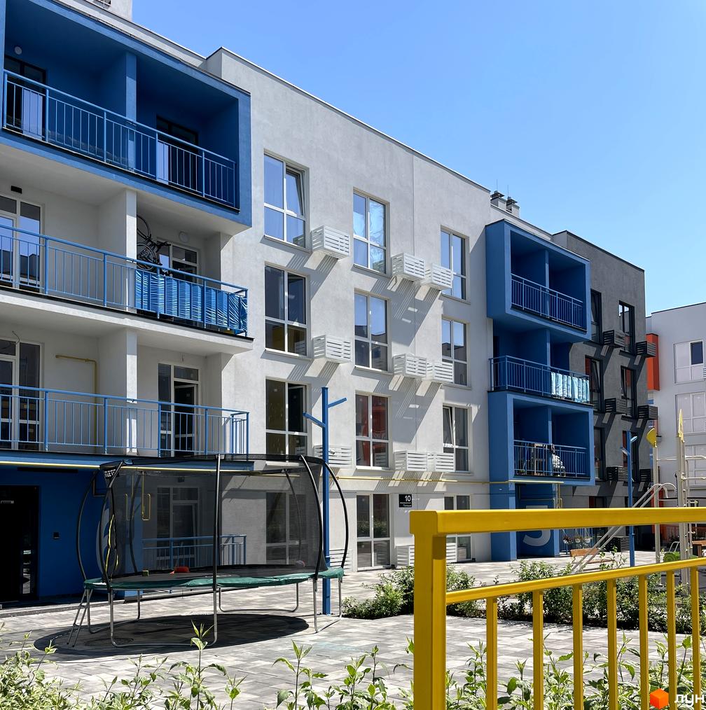 Житловий комплекс Art Misto: фасади будівель з синіми балконами та сірими елементами на тлі блакитного неба. Поруч дитячий майданчик з батутом та зелені насадження.