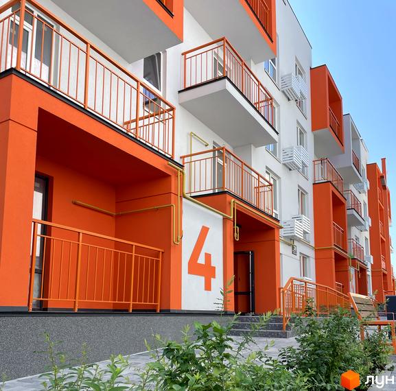 Фасад житлового комплексу Art Misto з яскравими помаранчевими елементами та балконами. Територія перед будинком з зеленими насадженнями та під'їздом.