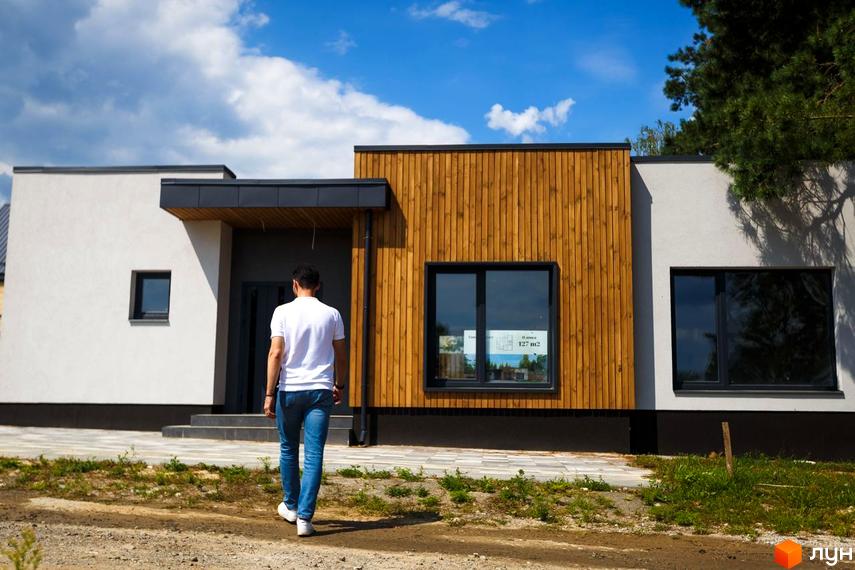 Одноповерховий будинок VISION з білим фасадом та дерев'яними панелями, темними вікнами. Перед будинком ґрунтова ділянка та вимощена доріжка.