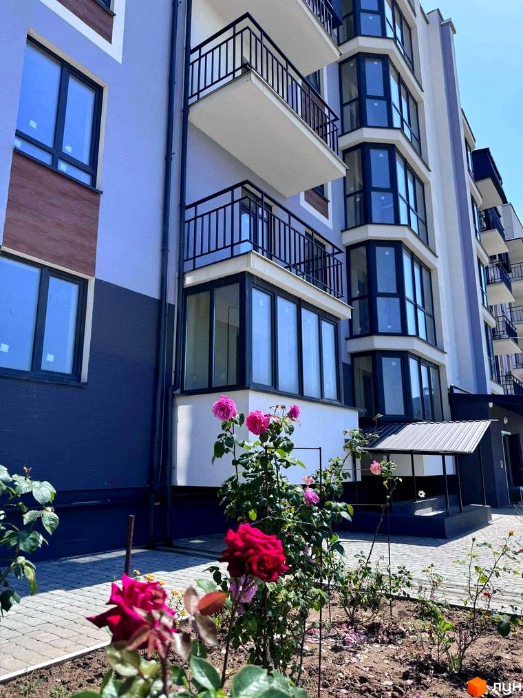 Зовнішній вигляд новобудови ЖК 9 Район з балконами та вікнами, прибудинкова територія з квітами та бруківкою.