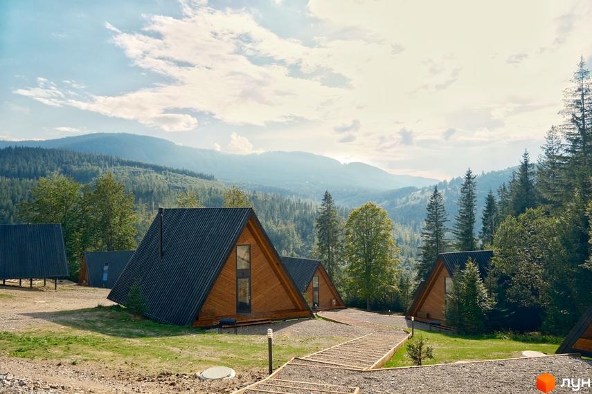 Комплекс відпочинку УЛІС: трикутні дерев'яні будинки з темними дахами на тлі гірського лісу. Територія комплексу з доріжками та зеленими насадженнями.