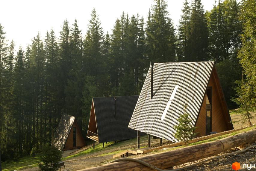 Комплекс відпочинку УЛІС: три трикутних будиночки на схилі гори серед ялинового лісу. Навколо зелена трава та гравійна доріжка.