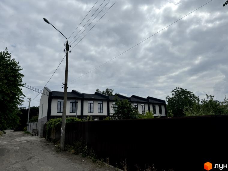 Таунхаус „Дивногорський“ з білими стінами та темним дахом на тлі сірого неба. Навколо зелені дерева та паркан.