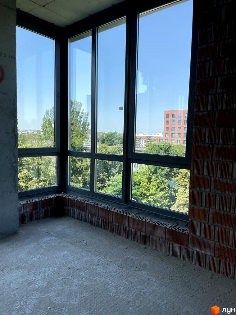 Вид з вікна на будівництво ЖК AcademHoff, що складається з цегляних стін та великих вікон з видом на зелені дерева та міські будівлі.