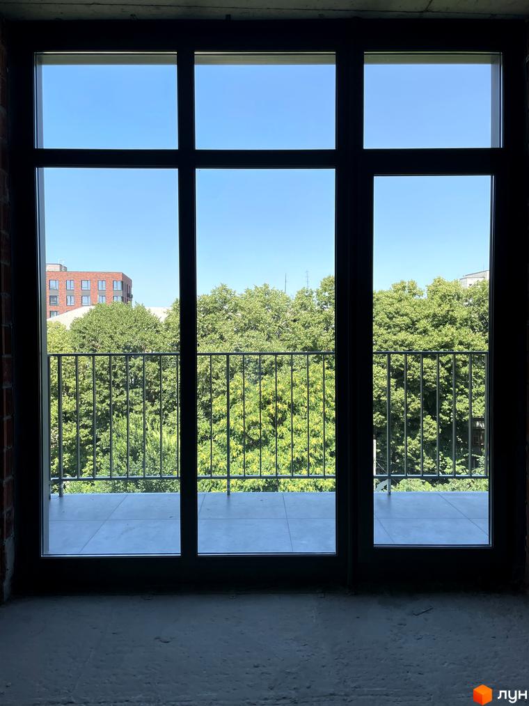 Вид з вікна на цегляний будинок та зелені дерева. Балкон з металевою огорожею перед вікном.