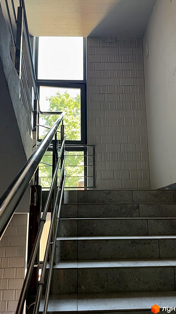 Сходи в житловому комплексі ЖК AcademHoff з металевими поручнями та великим вікном, через яке видно зелені дерева. Зовнішній вигляд будинку та прибудинкова територія.