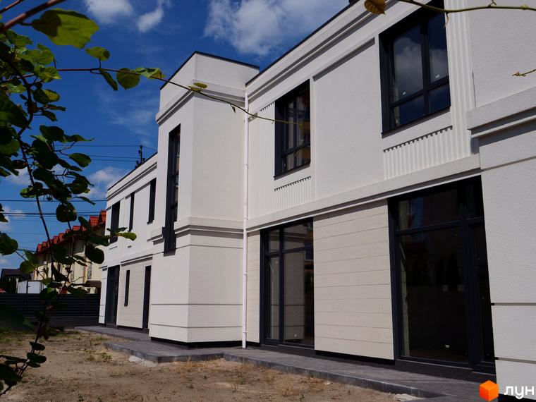 Біла будівля з великими вікнами на тлі блакитного неба. На передньому плані видно зелене листя, а поруч – прибудинкова територія.