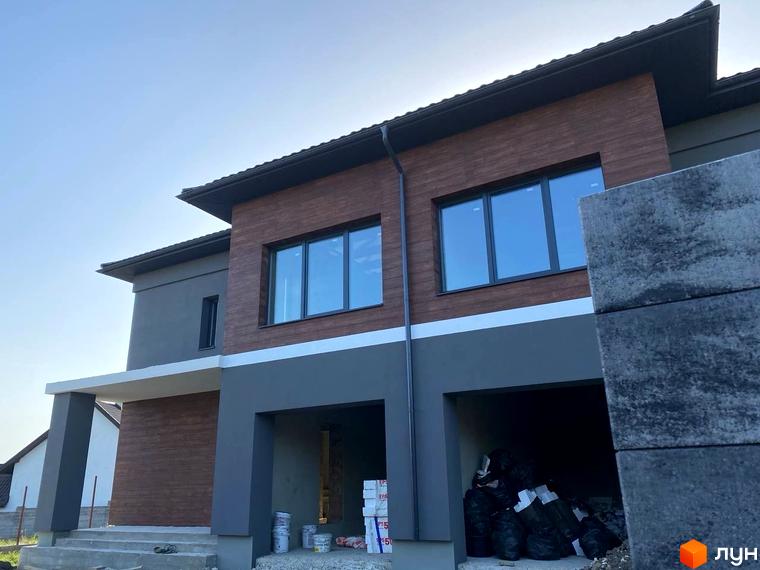 Фасад будинку з коричневою та сірою обробкою, великими вікнами та навісом над входом. Перед будинком розташовані сходи та будівельні матеріали.