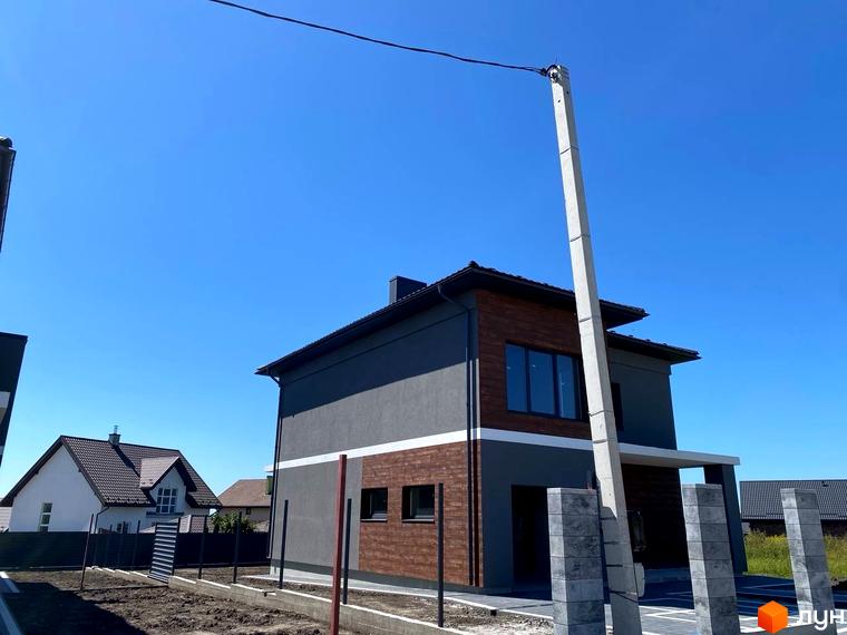 Сучасний двоповерховий будинок з сірою та коричневою обробкою фасаду, розташований на фоні блакитного неба. Поруч видно паркан та елементи благоустрою території.