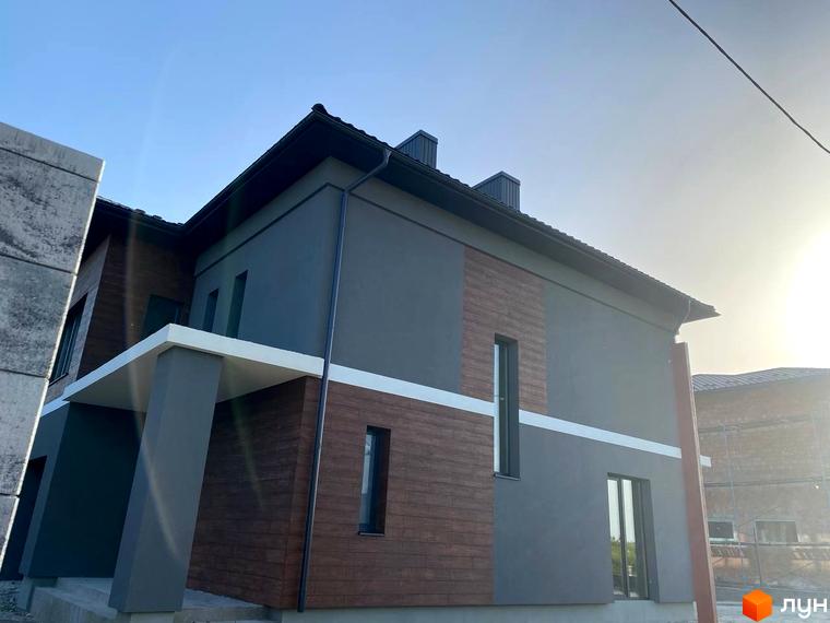 Фасад новобудови на вулиці Волошкова з сірим та дерев'яним оздобленням, що виглядає акуратно та сучасно. Навколо будинку видно елементи прибудинкової території.