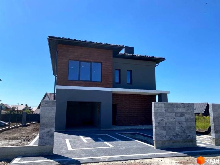 Сучасний двоповерховий будинок з коричневими дерев'яними вставками та сірим фасадом, розташований на бруківці з білими лініями.