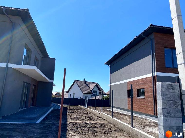 Зовнішній вигляд двоповерхових будинків з сірим фасадом та дерев'яними вставками на вулиці Волошкова. На прибудинковій території видно огорожу та ділянку з землею.