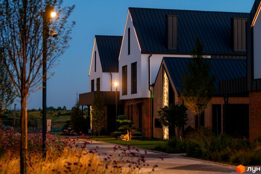 Вечірній вигляд Котеджного містечка LAGOM з будинками з білими фасадами та темними дахами. Освітлена доріжка та зелені насадження створюють затишну атмосферу.