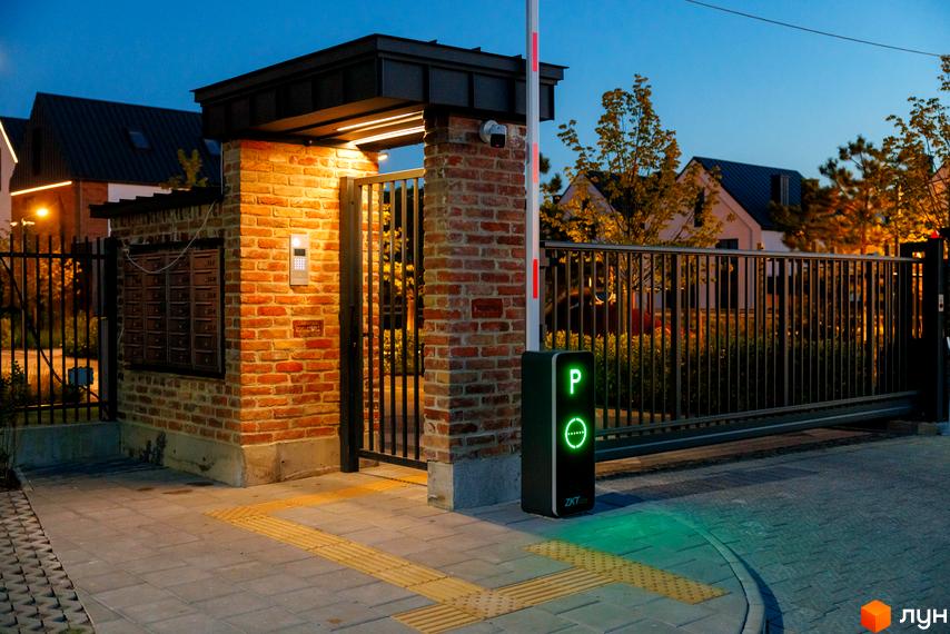 В'їзд до котеджного містечка КМ LAGOM з автоматичним шлагбаумом та домофоном. Огороджена територія з цегляною колоною та поштовими скриньками.