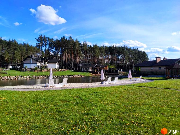 Зовнішній вигляд котеджного комплексу КМ Swisstown з прибудинковою територією та озером. На газоні розташовані шезлонги та парасольки.