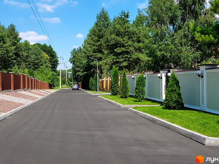 Котеджне містечко КМ Swisstown. Асфальтована дорога веде до будинків, обабіч якої розташовані цегляні паркани та зелені насадження. На території комплексу є доглянуті газони та декоративні туї.