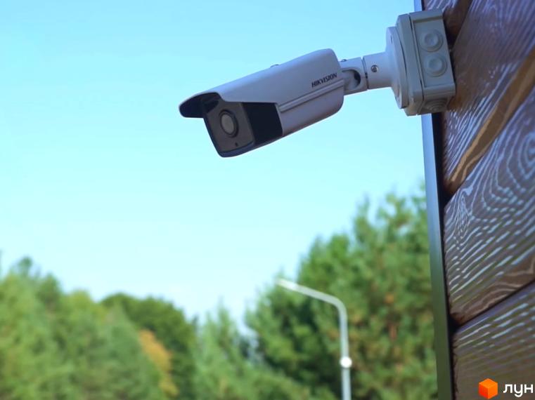 Зовнішній вигляд камери відеоспостереження на фасаді будинку житлового комплексу КМ Swisstown. Навколо зелені дерева та блакитне небо.