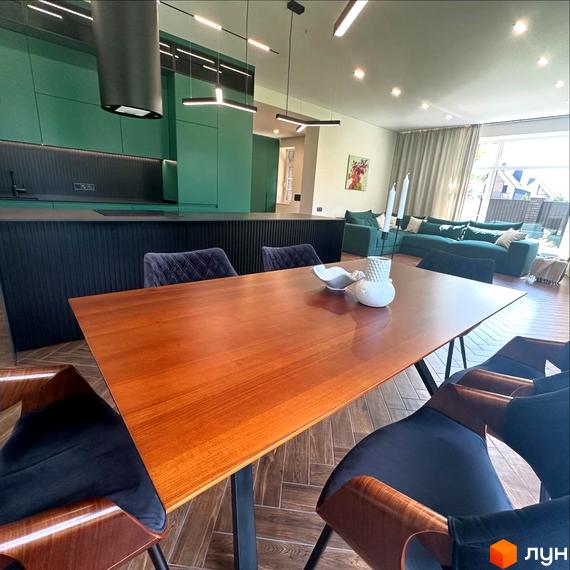 Просторий обідній стіл з дерев'яною стільницею та темно-синіми стільцями на тлі зеленої кухні та вітальні з диваном.