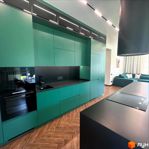 Сучасна кухня з зеленими фасадами та чорною стільницею. Простора вітальня з бірюзовим диваном видно на задньому плані.
