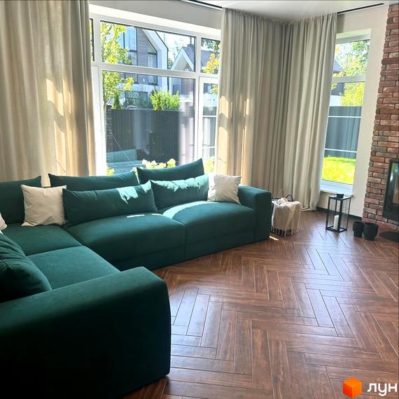 Простора вітальня з великими вікнами та зеленою кутовою софою в житловому комплексі Spacious HOME. Затишна атмосфера з цегляною стіною та видом на зелену ділянку.