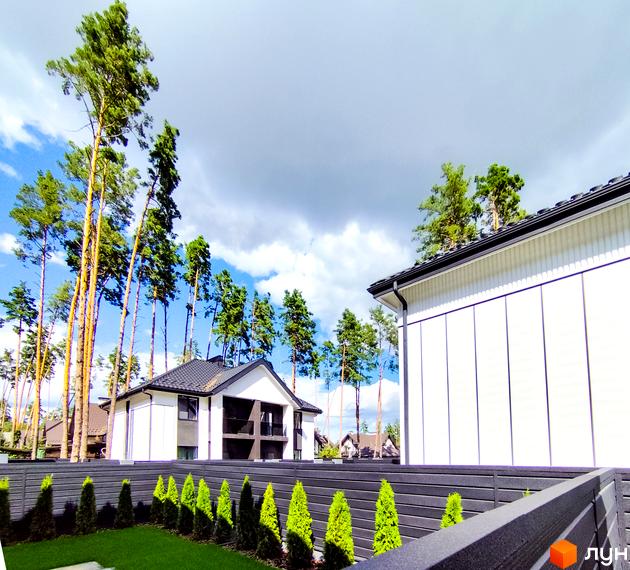 Білий будинок з темним дахом на тлі блакитного неба. Перед будинком розташована зелена галявина та огорожа з зеленими кущами.